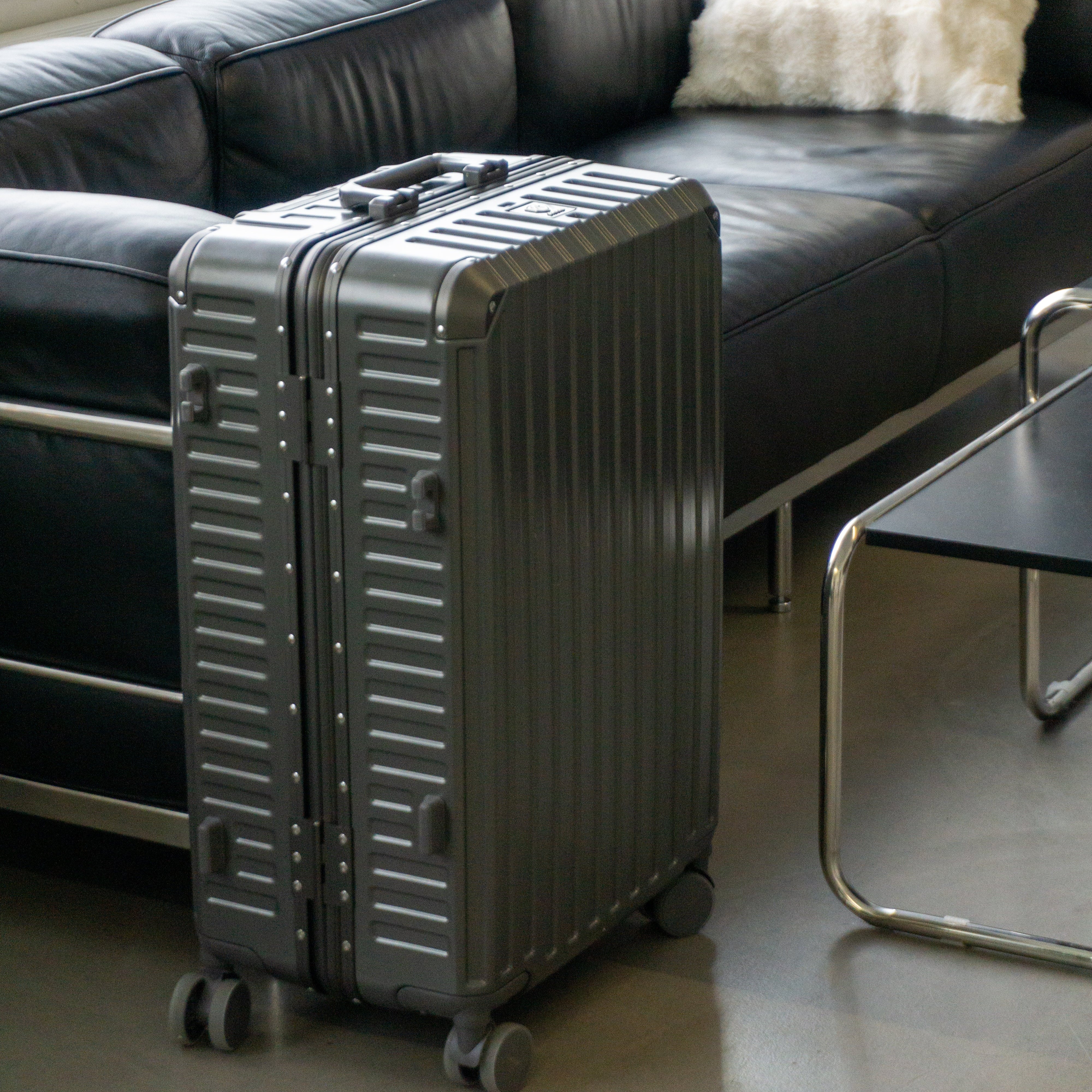Midnight grey luggage on a floor in a room with a black leather couch and white pillow.